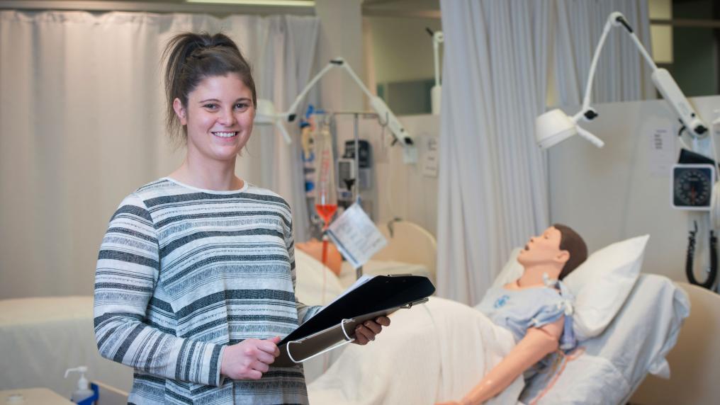 A woman standing next to hospital beds holding a chart and smiling at camera. A dummy patient lies in one of the beds.