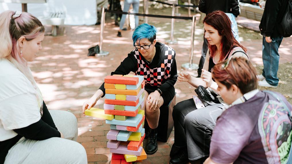 A group of students with bright hair play a block-stacking game in a sunny courtyard