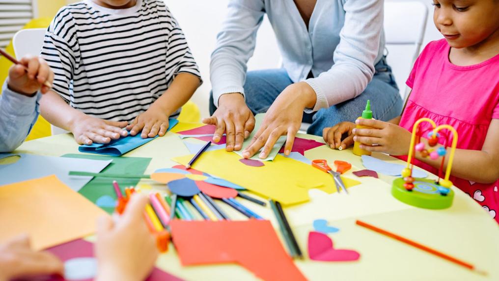 Children around a table with various pieces of paper and craft equipment. An adult is demonstrating how to fold a piece of paper. 