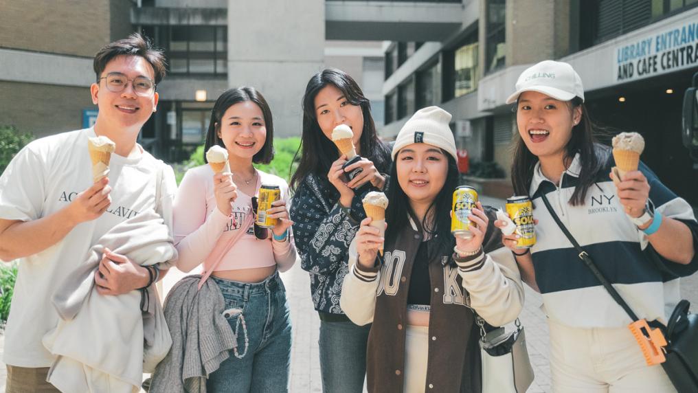 Students standing outside, holding an ice-cream and soft drink and smiling at camera.