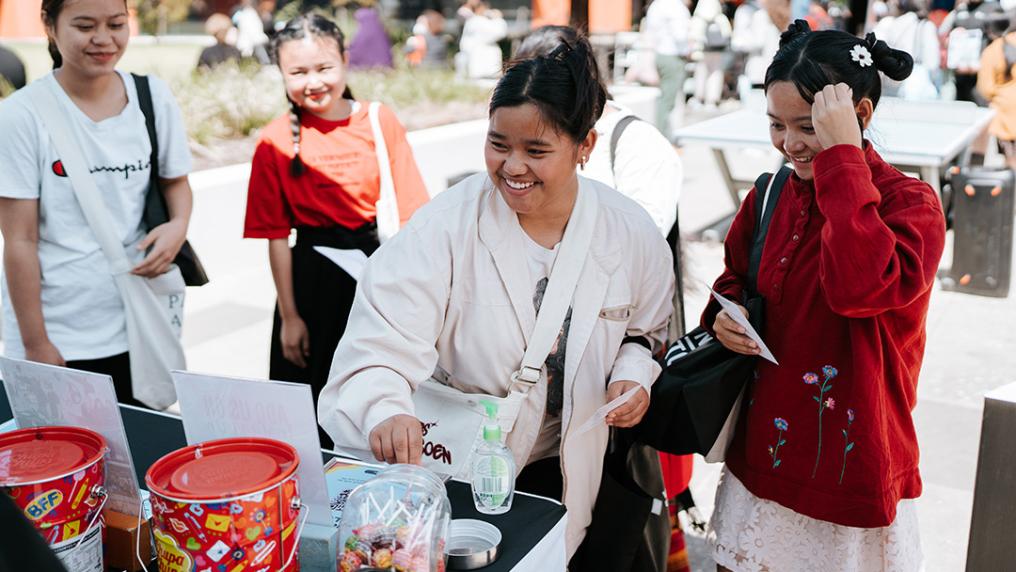 A group of young adults smile and help themselves to lollipops from a stall in a sunny courtyard