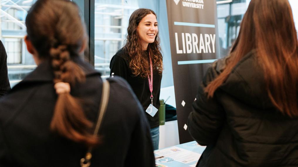 A person standing behind a stall next to a sign that says "Victoria University Library". People are gathering around the stall.