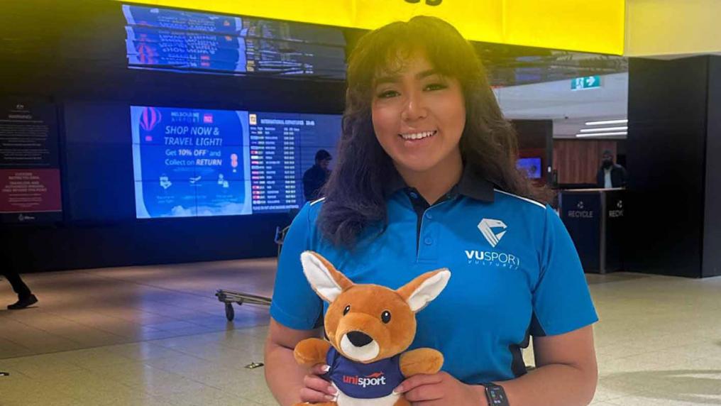 Stella Rose Lay standing in an airport wearing a VU Sport shirt holding a plush kangaroo with the UniSport logo.