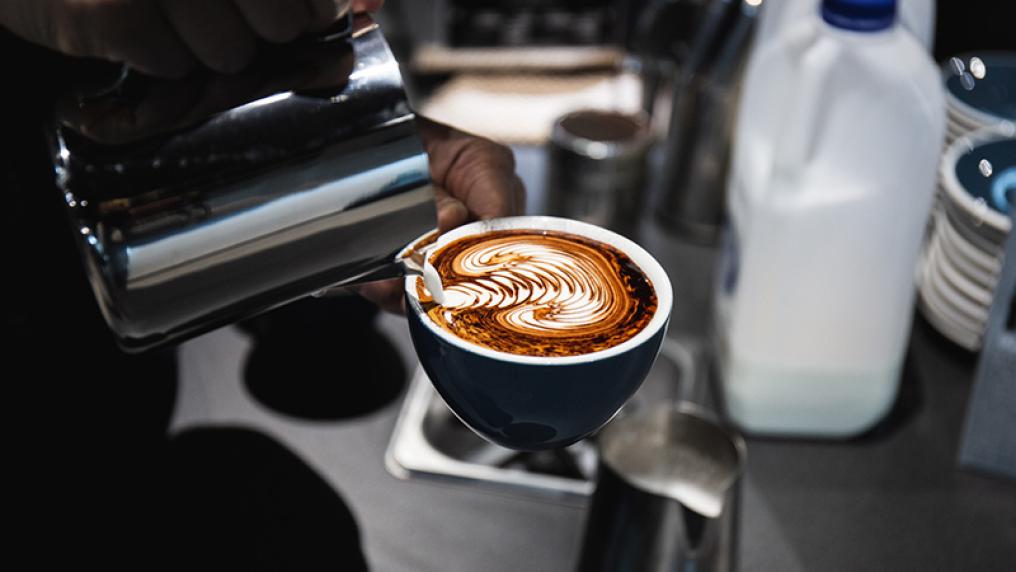 A milk jug is used to create a detailed pattern in milk on a coffee