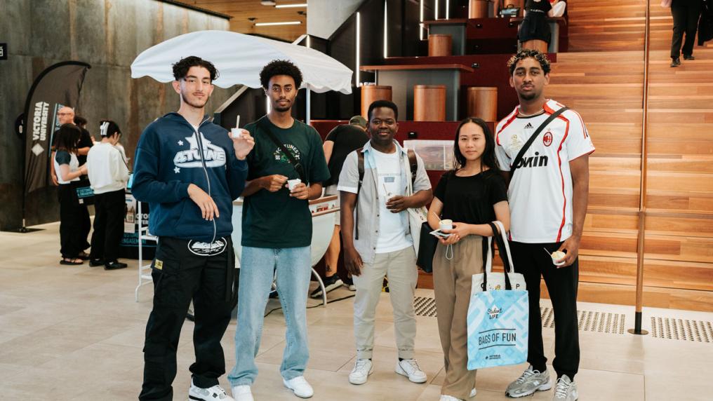 A group of 5 students inside City Campus, looking at the camera. Most of them are holding small cups of ice cream. Behind them is a gelato stall with a white umbrella above it.