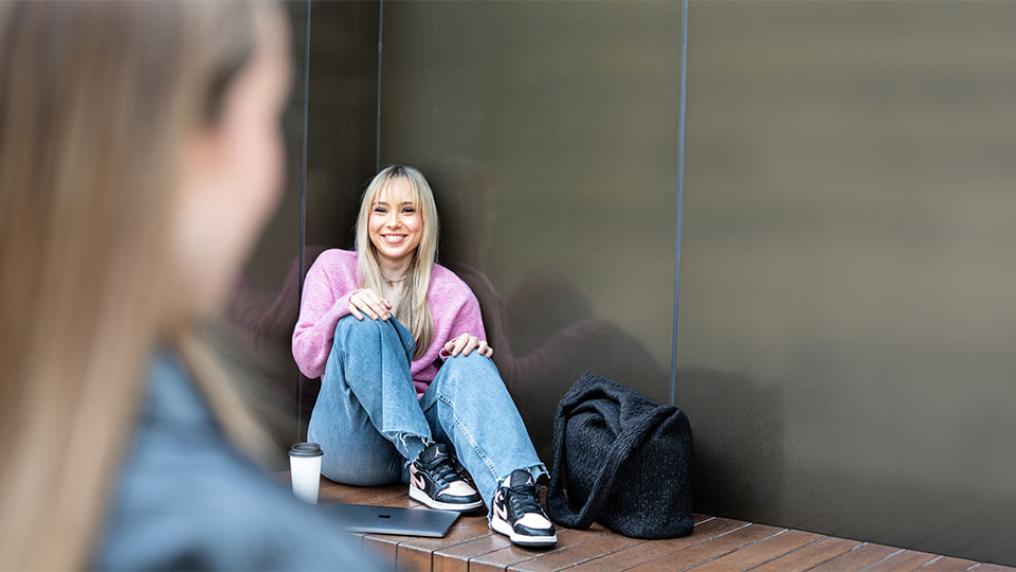 A VU student with long blonde hair, wearing jeans and a pink jumper sits against a wall with a black backpack placed next to her.