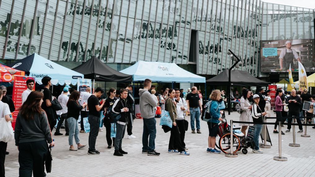A crowd of people outside P Block at Footscray Park Campus. Around them are a series of outdoor stalls and flags from different companies.