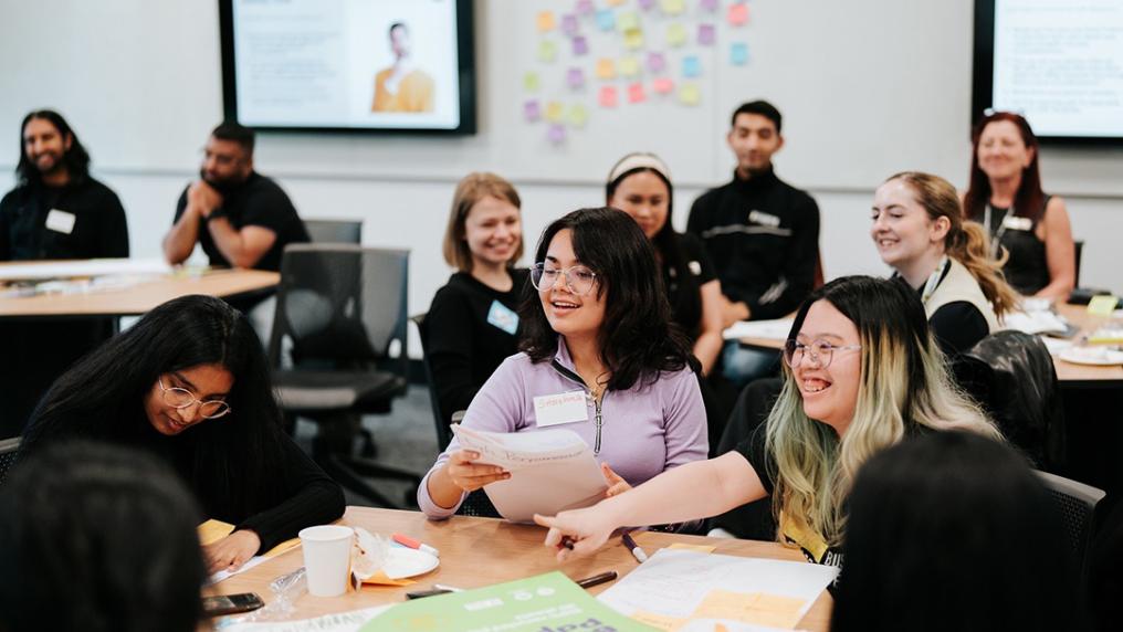 A group of young people in semi-formal clothes laugh around tables while attending a workshop