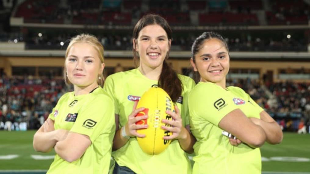 Three girls in umpire uniform standing on a field. One of them is holding an AFL ball.