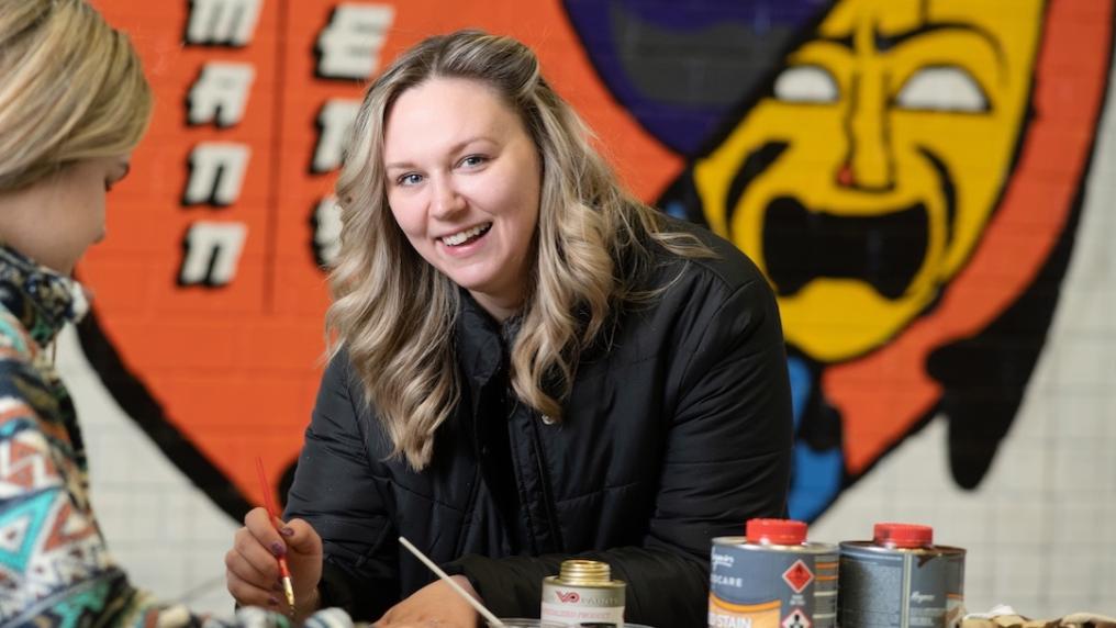Chloe painting while smiling at the camera. Behind Chloe is a stylised mural painted on the wall 
