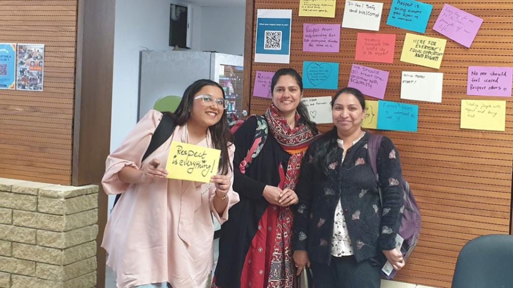 Three students standing in front of a wood panel wall. The wall contains pieces of paper with messages about respect.