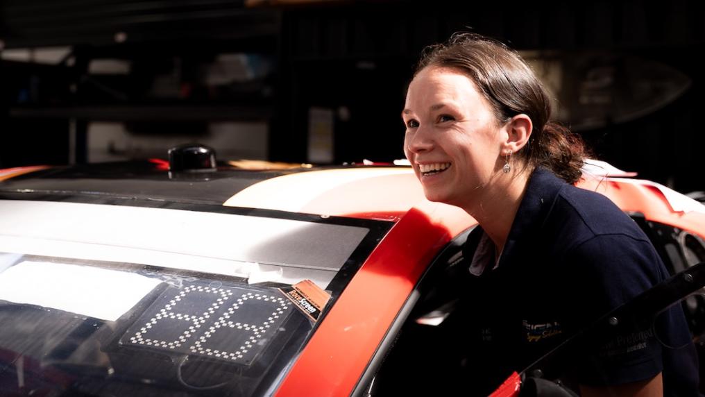 Jordyn McConchie next to a red racing car. The car has been modified with a number display on the front window for racing.