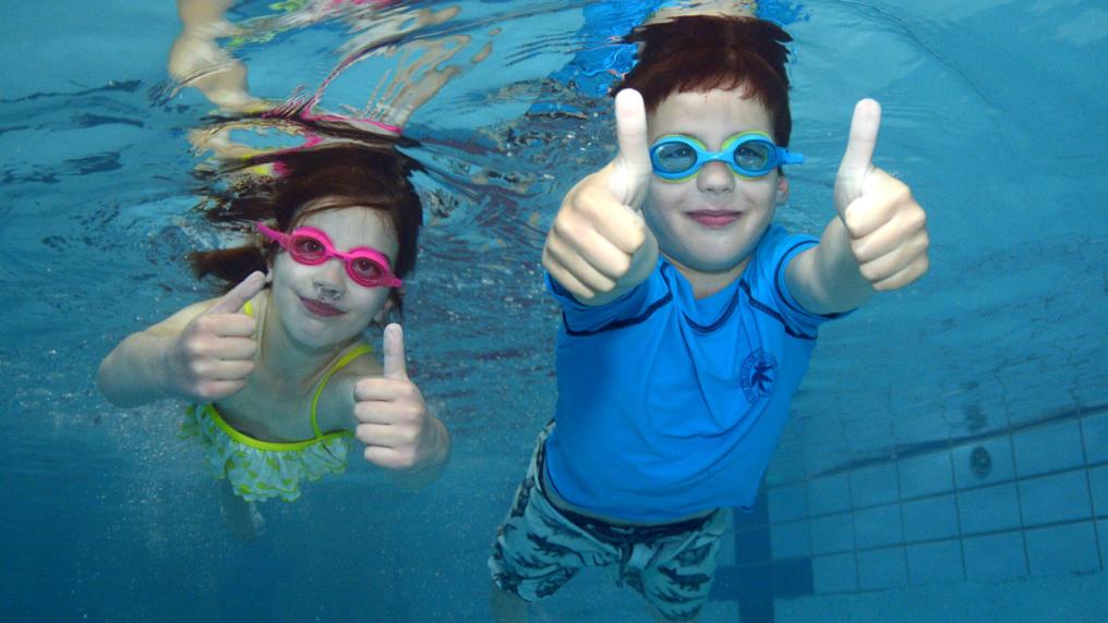 Two children swimming underwater. Both are holding their thumbs up at the camera.