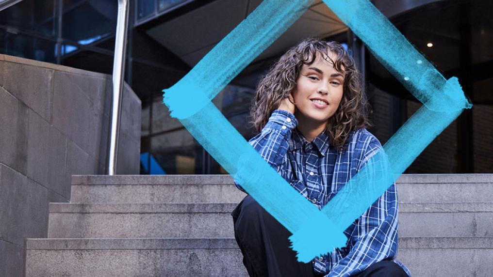 A student with curly hair in a blue plaid shirt sits on stairs with the VU blue diamond in the foreground.