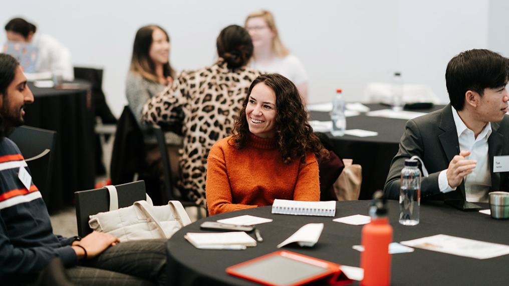 A workshop with a group of animated students around tables with papers and laptops
