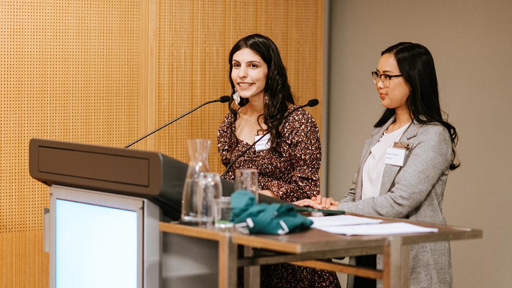 Two young women in semi-professional dress stand in front of microphones at a podium