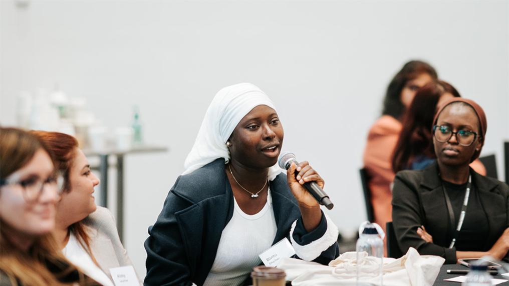 A woman sits at a table within a conference, talking into a microphone.
