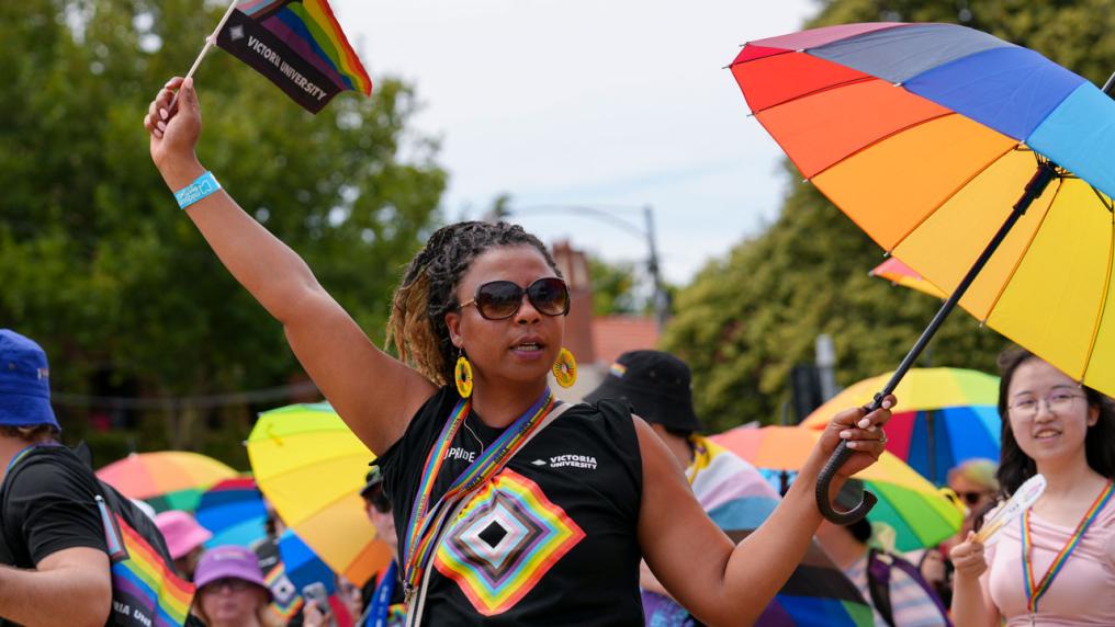 A person holding a pride umbrella and flag outside. They are wearing a pride shirt with the VU logo on it. Other people are standing around them with pride umbrellas.