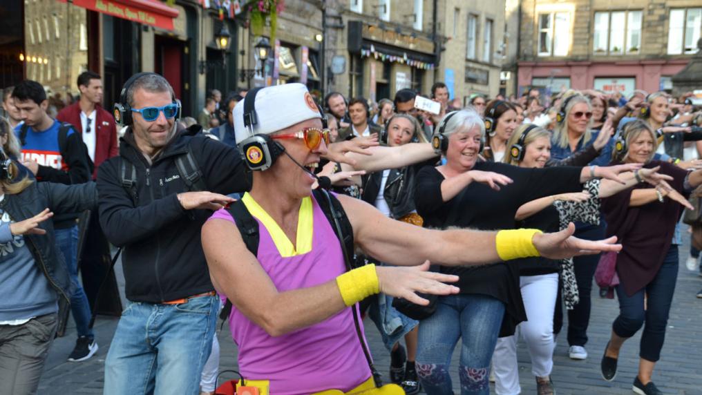 A large group of people outside on the street with headphones on. At the front is a person wearing bright-coloured clothes, glasses and wristguards. They're all coordinating the same dance move.