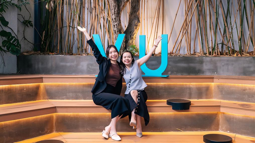 Two women in professional dress smile in front of a VU sign and bamboo background