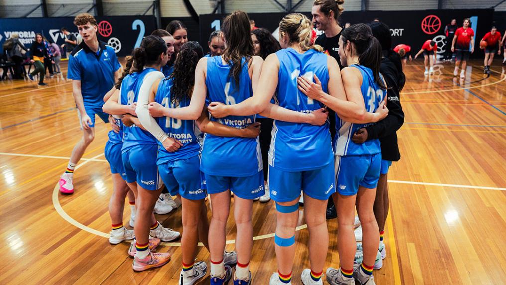 A group of people with long hair in pony tails wearing VU basketball uniforms, rainbow socks and sneakers huddle on a basketball court