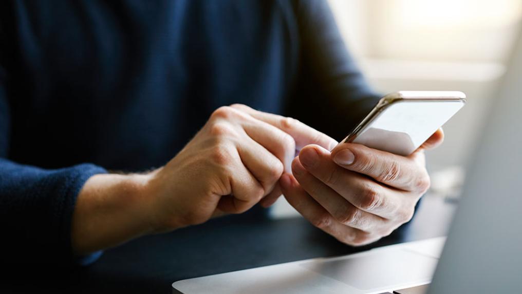 Hand holding mobile phone in front of a laptop.