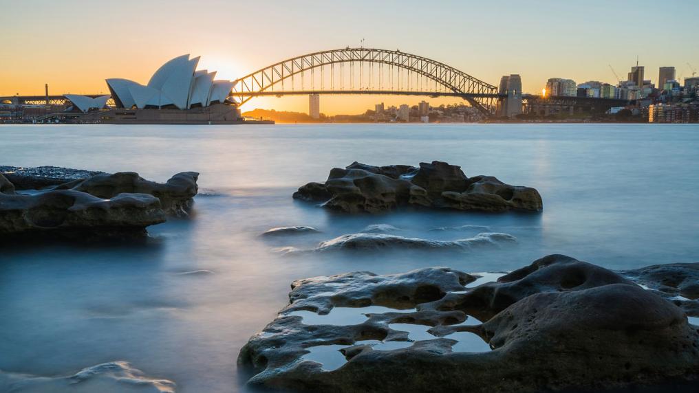 A photo of Sydney at sunset. The Sydney Opera House and Sydney Harbour Bridge can be seen in the distance.