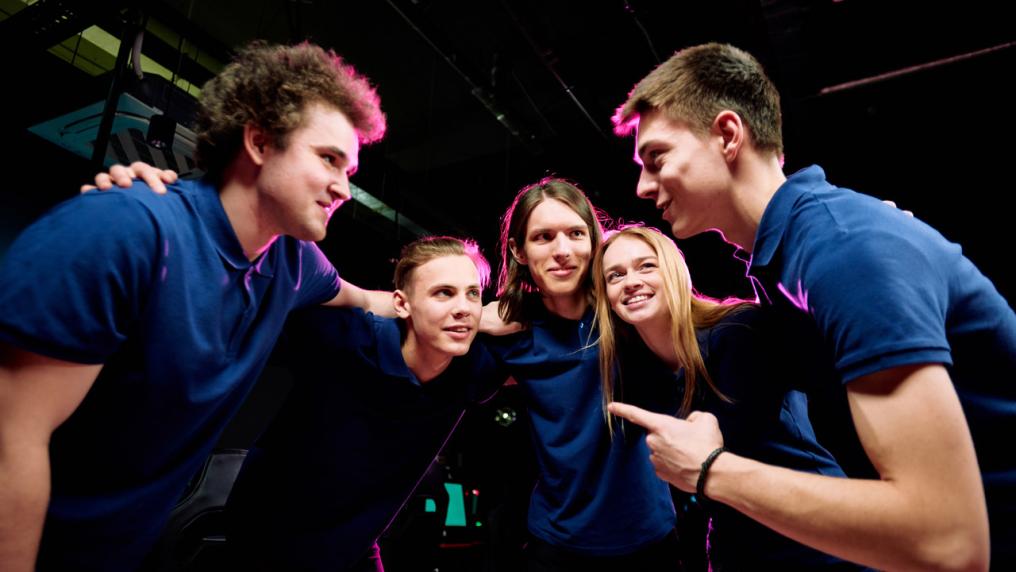 A group of students in an arcade huddling together. One person is speaking and pointing, the rest of the students are looking at him.