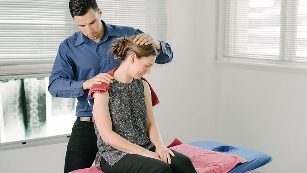 A man adjusts a woman's neck in a clinical setting with massage table and x-rays