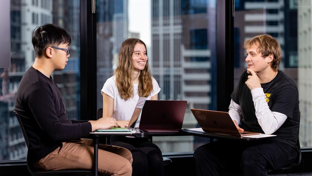 3 students discuss at a table with laptops, in room with a city view