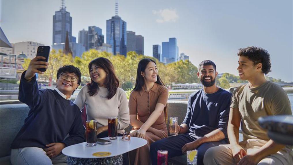 A group of international students smile on a bridge over a wide river, in front of a modern city skyline