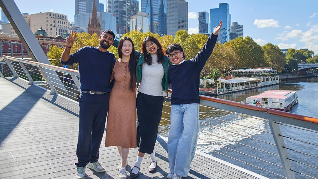 A group of international students smile on a bridge over a wide river, in front of a modern city skyline