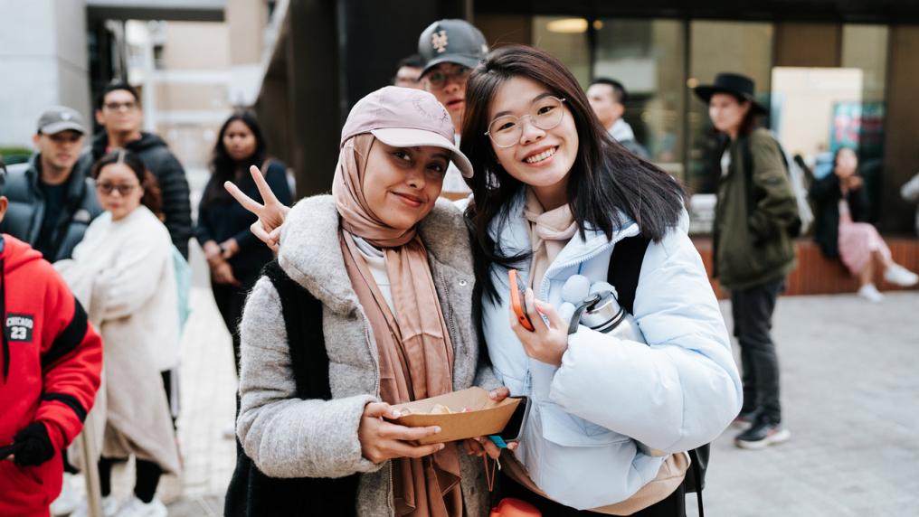 Two students standing outside and smiling at the camera. One student is holding a food carton. Behind them are groups of students walking around.