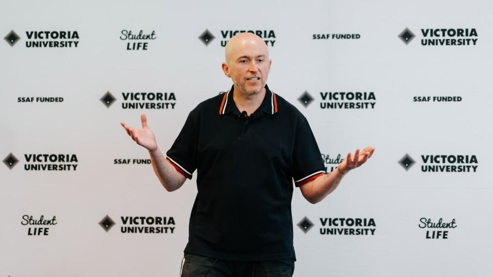 A person speaking at an event. They are standing in front of a wall with Victoria University and Student Life logos.