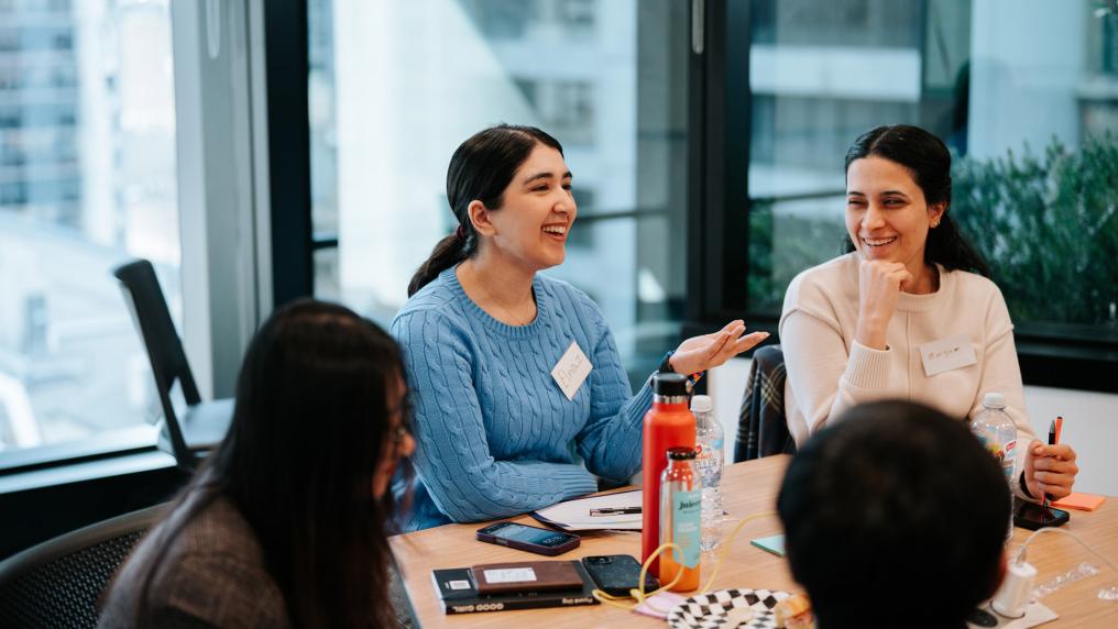 Students at a table. One is responding to a teacher. Another student is looking at the speaking student.