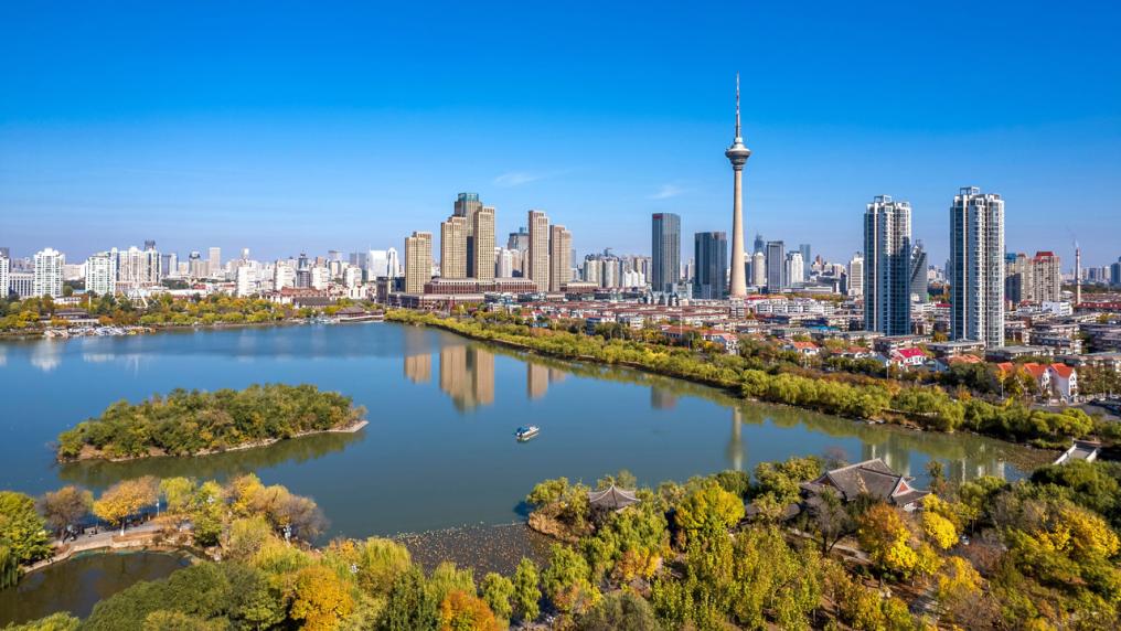 A photo of Shenyang, China. Modern skyscapers and a tower can be seen. Closer to the camera are lush green trees and a small island in a river.