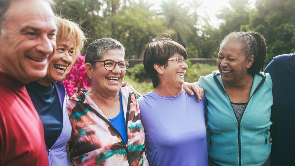 A group of senior people standing outside in a park, arms over each others' shoulders.