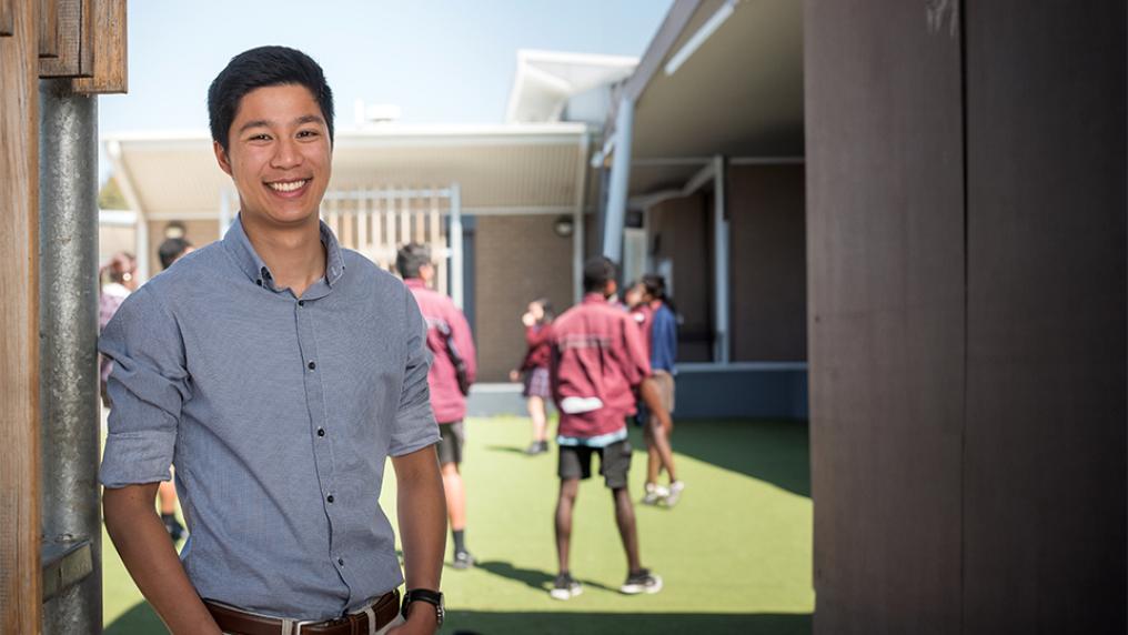 A man in a blue shirt poses at a high school, with kids playing sport in the background.