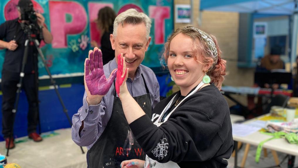 Vice Chancellor Adam Shoemaker and a student showing their hands to the camera. Their hands are covered in paint. Behind them is a painted mural with the word "RESPECT".