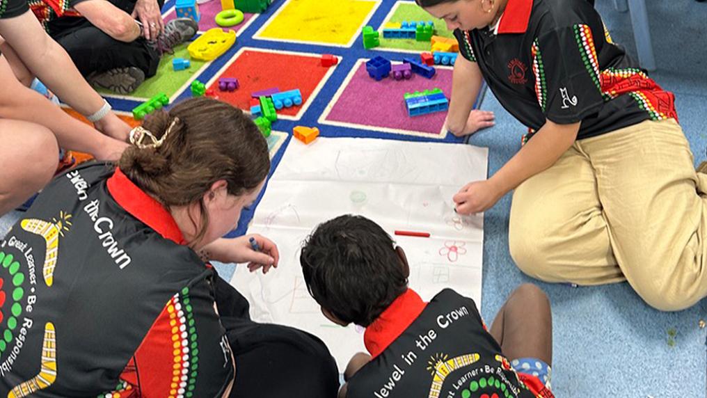 A group of young people wearing t-shirts with First Nations patterns draw on a classroom floor