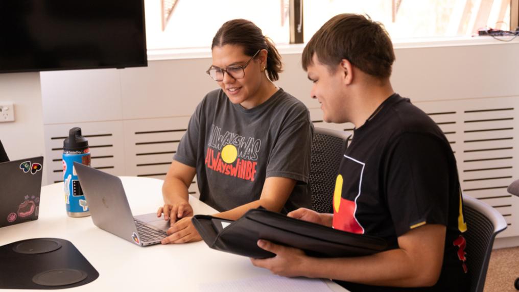 Two students wearing t-shirts with Australian Aboriginal flag, engaged in discussion and looking at laptop.