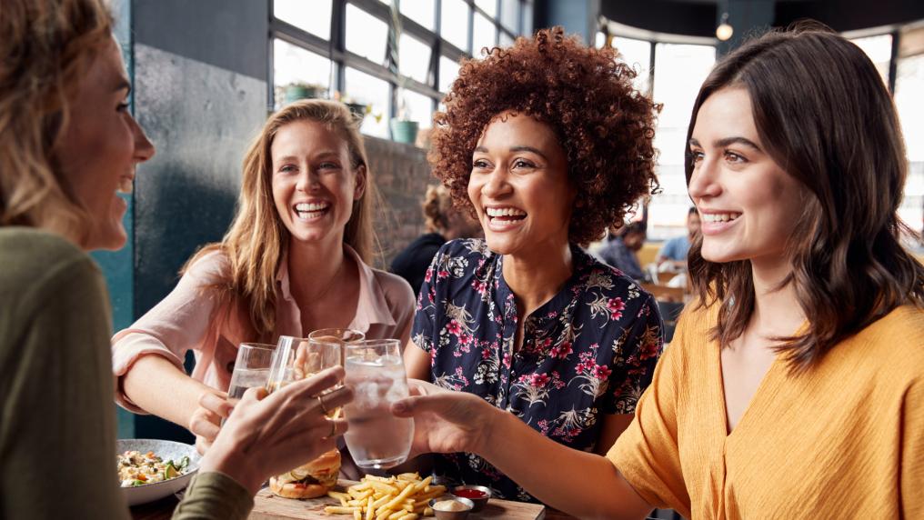 A group of women inside a restaurant smiling and tapping their drink glasses together.