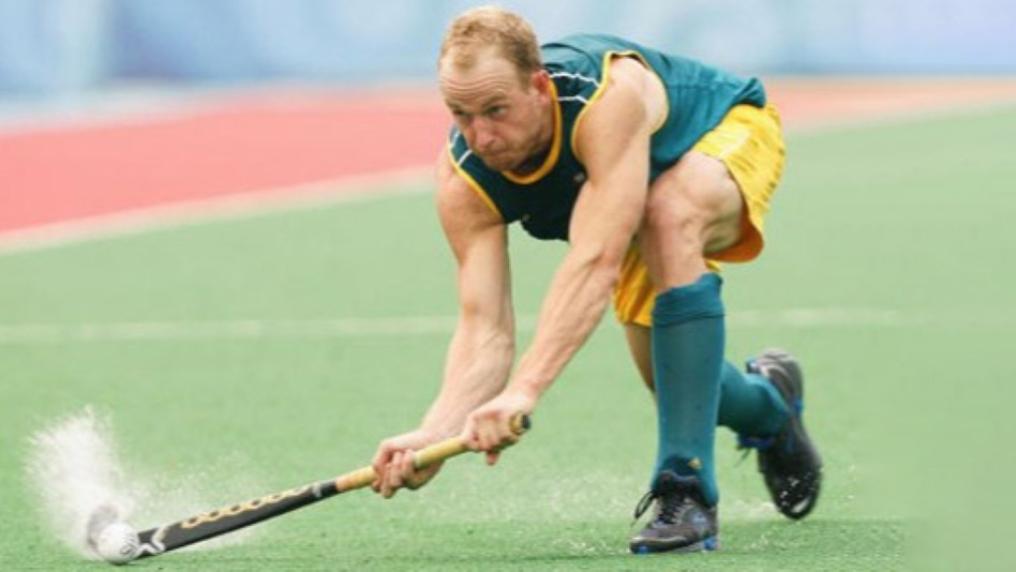 Male hockey player crouching on playing field hitting ball with hockey stick. 