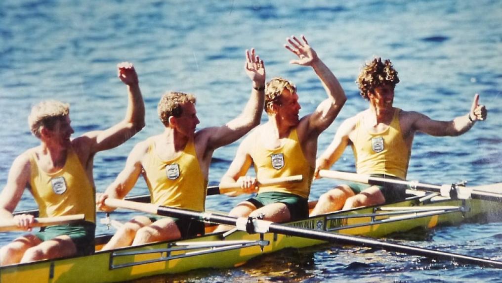 Four male rowers in rowing boat on river, waving and smiling. 