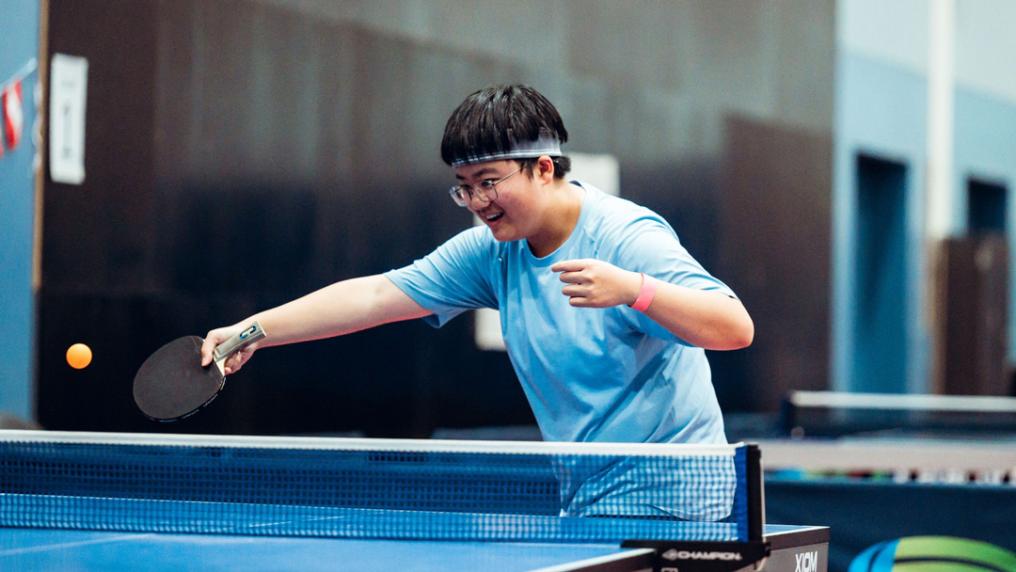 A young person in a VU blue t-shirt smiles while playing table tennis
