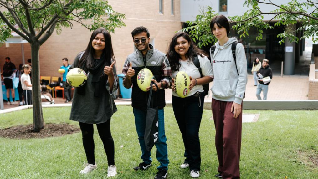 Four young adults with backpacks & casual clothes smile and pose in a grassy courtyard