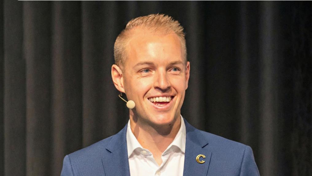 Smiling man in suit with headset speaking with black curtains in background. 