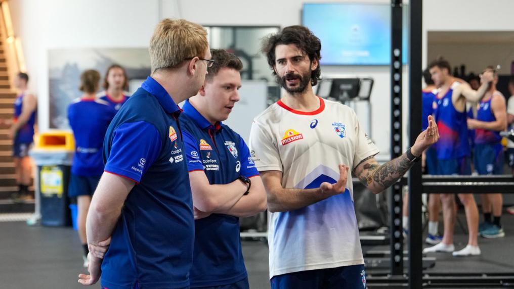 2 Western Bulldogs AFL players in a gym listening to one person is directing them.