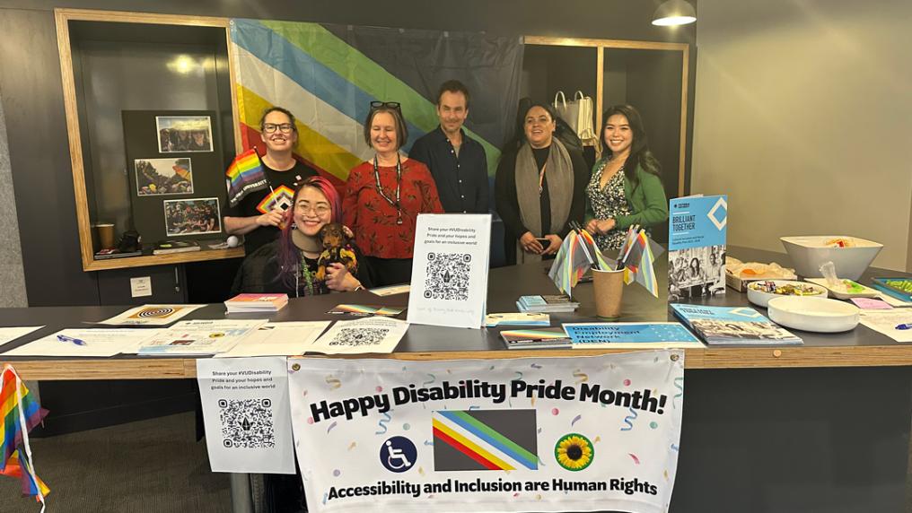 Group of VU staff members gathered around Disability Pride Month stall. There is a large Disabiliiy Pride Flag behind the group. On display in front of the group is a banner with text ' Happy Disability Pride Month' with the wheelchair disability logo, the Sunflower Hidden disability logo and text 'Accessibility and Inclusion are Human Rights'.