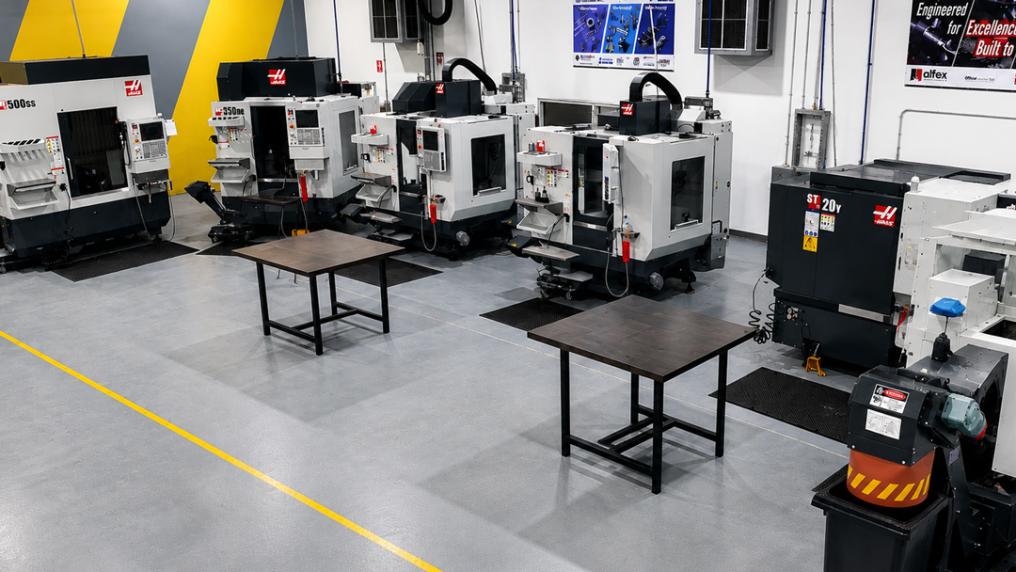 Several large automated machine tools sitting in an industrial-style room with small tables. 